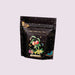 A stand-up pouch of Magic Hour Cosmic Carven Iced Teas on a soft pink background. The packaging is black with a starry night design, gold decorative borders, and an illustration of peach, fig, and yellow flowers.